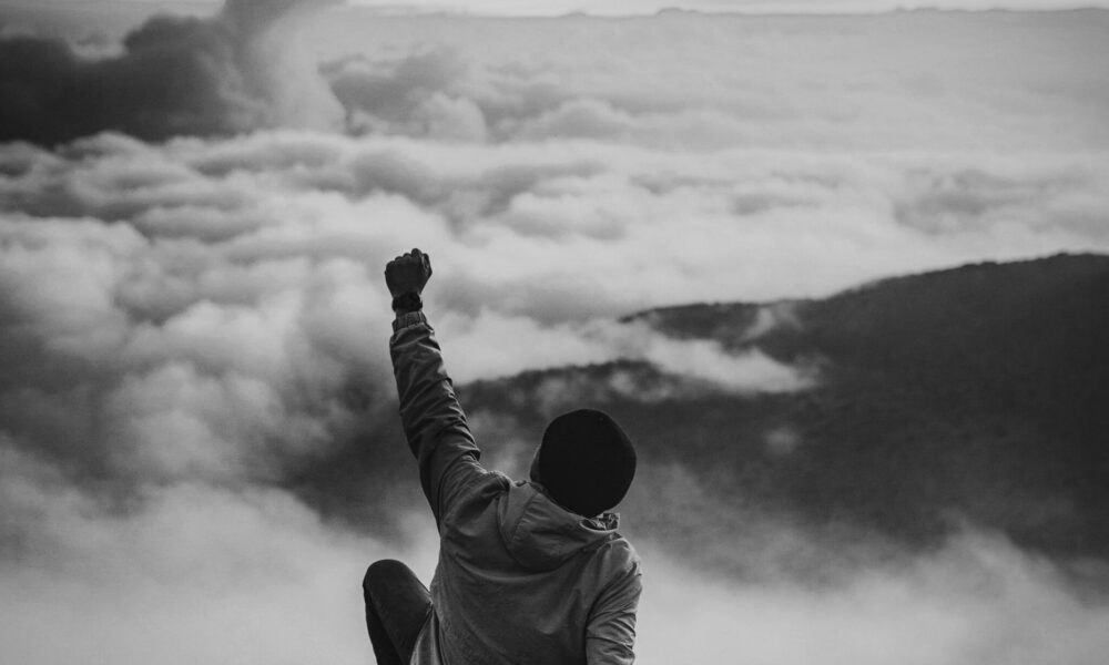 Mann sitzt auf Klippe mit erhobenem Arm und Blick auf die Wolken