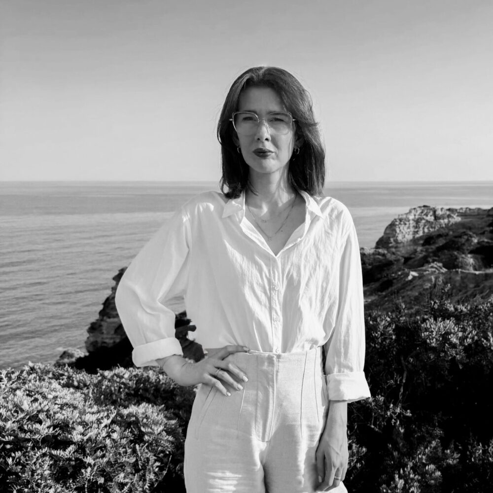 eine Frau steht mit dem rechten Arm an der Hüfte vor einer Klippe am Meer und schaut in die Kamerarichtung
